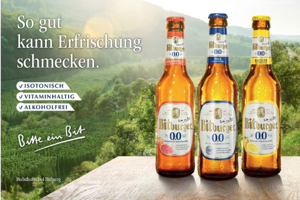 3 Bitburger Bierflaschen mit weißem Schriftzug vor grüner Landschaft