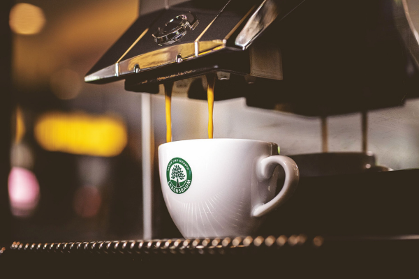 Weiße Cappucinotasse mit grünem Logo unter Kaffeevollautomat